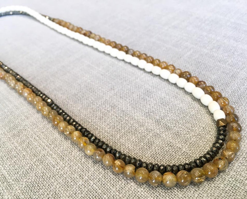 top view of gemstone bead necklace comprising white conch shell, dark grey pyrite and neutral rutilated quartz